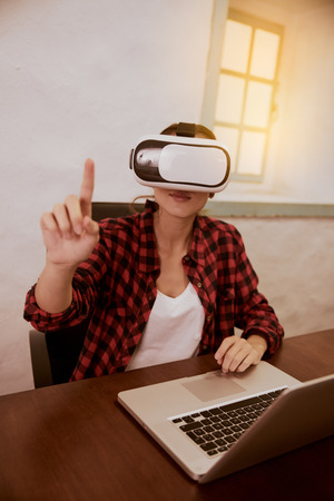 Young woman with a 3d viewer to her eyes sitting with her laptop facing the camera while holding her right finger upの写真素材