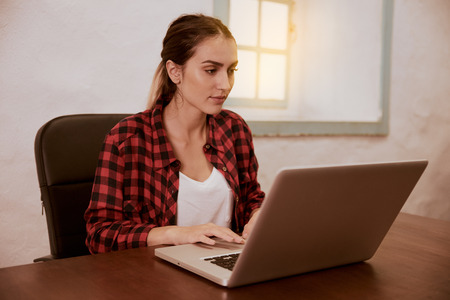Concentrating young lady typing on her laptop while sitting on an office chair at a desk with a white wall behind herの写真素材