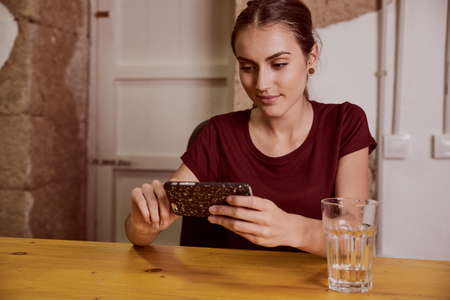 Cute young woman with a red top and her hair tied back sitting at a desk looking at her cell phone with a knowing smileの写真素材