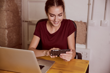 Beautiful young woman with a red top sitting at a desk smiling at her cell phone with her laptop open next to herの写真素材