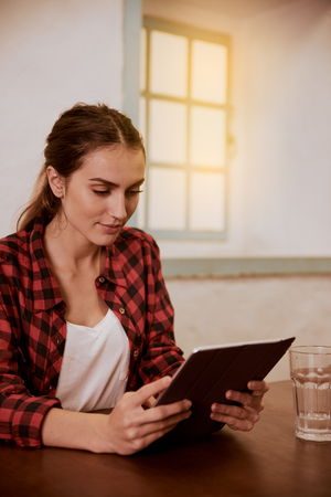 Young woman holding her tablet with both hands while sitting at a desk with a glass of water and a white wall behind herの写真素材