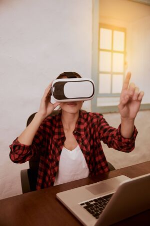Young woman with a 3d viewer to her eyes sitting with her laptop facing the camera while holding her left finger upの写真素材