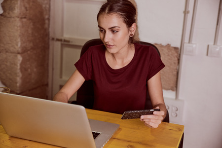 Stunning young lady with a red top sitting at a desk smiling at looking at her laptop with her cellphone on one handの写真素材