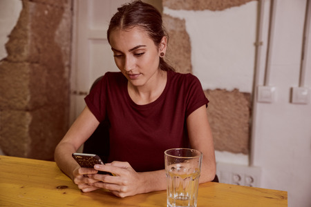 Cheerful young woman with a red top and her hair tied back sitting at a desk looking at her cell phone with interestの写真素材