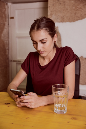 Beautiful young woman with a red top and her hair tied back sitting at a desk typing a message on her cell phoneの写真素材