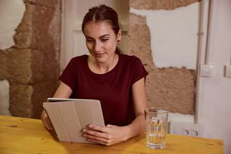 Beautiful young woman with a red top and her hair tied back sitting at a desk looking at her tablet with a smileの写真素材