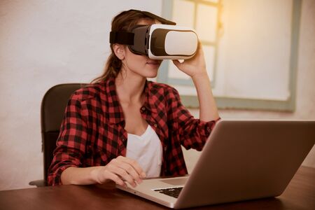 Beautiful young woman holding a 3d viewer to her eyes while sitting at a desk with her laptop in front of herの写真素材