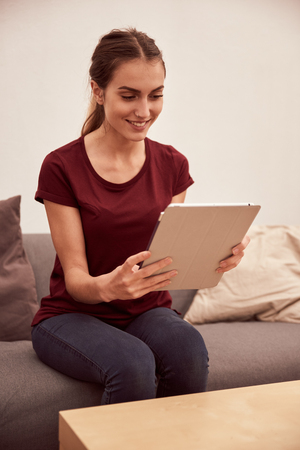 Beautiful young lady holding her tablet in both hands while sitting on a grey couch with scatter cushions and white light from behindの写真素材