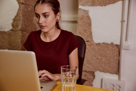 Beautiful young woman with a red top and her hair tied back sitting at a desk with her laptop and a glass of waterの写真素材