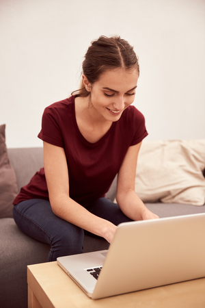 Relaxing young woman sitting on a grey couch with cushions typing on her laptop with bright white light from behind herの写真素材