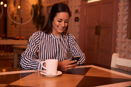 Beautiful young woman wearing a striped shirt in a restaurant reading message on her cellphone with a mug of coffee to her rightの写真素材