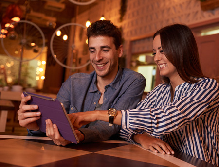 Happily laughing young couple sitting very relaxed at a table in a restaurant while pointing and looking at a tabletの写真素材