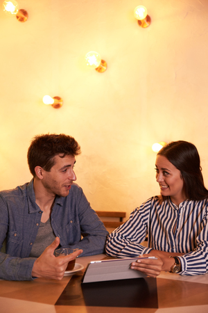 Interested young couple looking at each other while sitting in a brightly lit restaurant with a tablet between themの写真素材