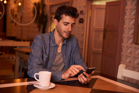 Thinking young man wearing a denim shirt in a restaurant typing on his cellphone with a mug of coffee to his rightの写真素材