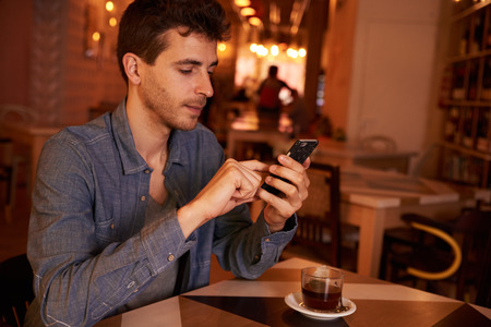 Charming millenial sitting at table in restaurant texting with concentration on his cell phone with a cup of tea on the tableの写真素材