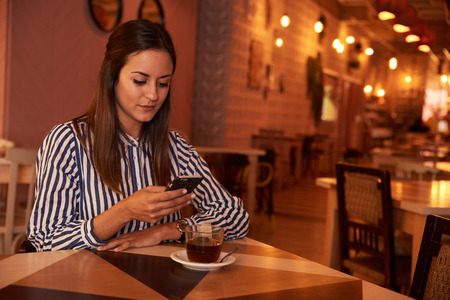 Thoughtful millenial sitting in a restaurant reading a message on her cellphone with a cup of tea on the table in front of herの写真素材