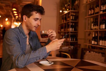 Charming male millenial sitting at table in restaurant reading a message on his cell phone while holding his tea upの写真素材