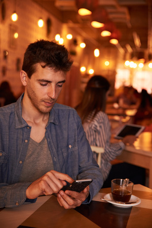Unshaven millennial sitting in restaurant with a cup of tea while texting with a longhaired girl sitting behind him with a tabletの写真素材