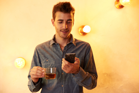 Cute young man reading a message on his cellphone with a cup of tea while standing in front of a brightly lit wallの写真素材