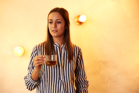 Woman looking and smiling into the distance while holding a cup of tea as she stands in front of a brightly lit wallの写真素材
