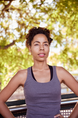 Fit young woman standing with her hands on her hips looking dreamily into nothingness with the sun shining through the leaves of a treeの写真素材