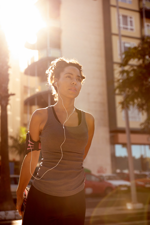 Female jogger with a music player on the side of a street with buildings and cars behind her and bright reflective sunshineの写真素材