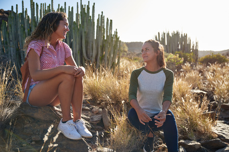 Two girl friends sitting on some rocks enjoying a moment of intimate chatter with cacti behind them in the late afternoon sunの写真素材