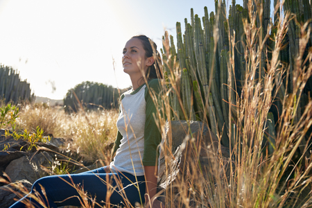 Lovely girl sitting on a rock in front of a large rain flower cactus on a grassy hill looking dreamily ahead of her in sunlightの写真素材