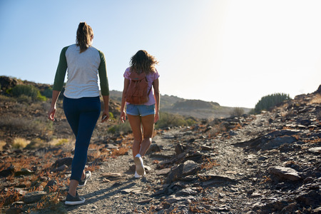 Two young ladies strolling up a steep hill wearing casual clothing and a backpack with their arms beside them in the afternoon sunの写真素材