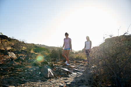 Two girls strolling down a rocky hill wearing casual clothing with their arms beside them in bright reflective afternoon sunの写真素材