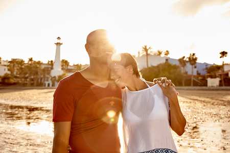 Laughing embracing adult couple on the beach with their arms around each other and hands interlocked, her head on his shoulder in very bright sunshineの写真素材