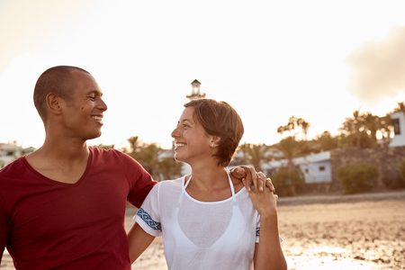Loving adult couple on the beach with their arms around each other and eyes locked together with a clear white sky behind themの写真素材