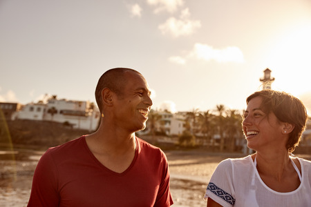 Loving adult couple walking on the beach while looking at each other lovingly with a light tower behind them and bright reflective white skyの写真素材