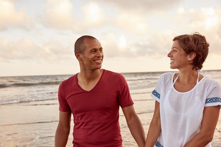 Charming adult couple in love looking at each other on the shore while enjoying the breaking waves of the sea behind themの写真素材