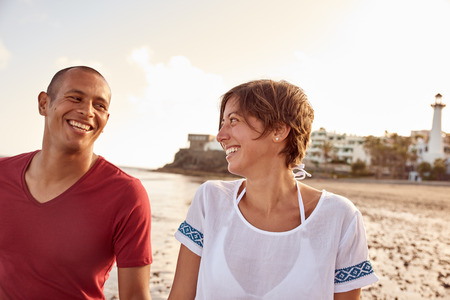 Loving couple laughing with joy while looking at each other while taking a relaxing walk on the beach with a clear white sky behind themの写真素材