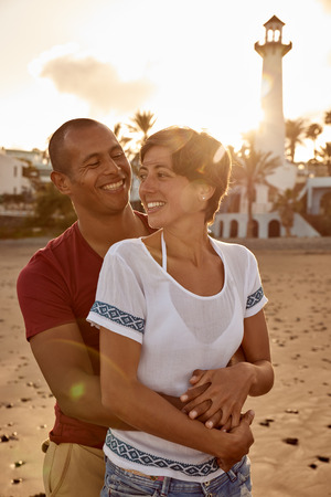 Beautiful couple in love on the beach in an embrace enjoying the last rays of the sun as it sets behind themの写真素材