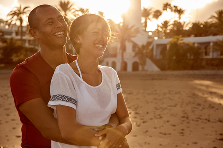 Embracing adult couple in front of a lighthouse in a loving embrace laughing with love while the sun is setting behind themの写真素材