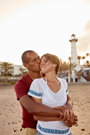 Loving adult couple on the beach in an embrace as he gives her a peck on the cheek while he holds her from behindの写真素材