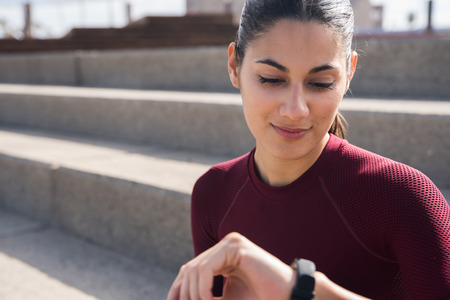 Young pretty female looking at smartwatch wearing maroon fitness shirt with hair tied backの写真素材