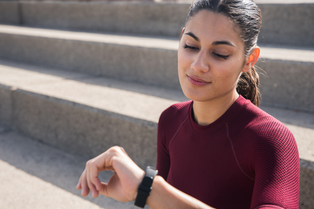 Young beautiful girl looking at sport watch wearing maroon sporty shirt with hair tied backの写真素材