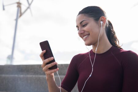 Young pretty woman looking at phone and smiling while wearing ear phones and a maroon shirtの写真素材