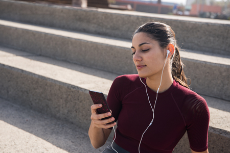 Young attractive female with cell phone on stairs wearing ear phones and a maroon sport shirtの写真素材