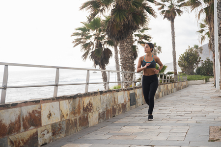 A young woman running along the promenade in black tights and a sports top with the palm trees in the background against a white skyの写真素材