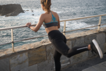 Back view of a woman exercising by the sea wearing black tights and tank top with her leg stretched backの写真素材