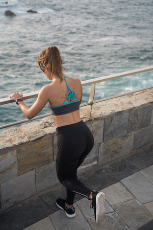 A sporty woman looks down at the sea while using a railing to do her exercisesの写真素材