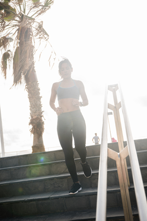 A young woman in bright sunlight running down the stairs with the white sky and a palm tree in the backgroundの写真素材