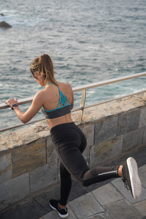 A slim young woman exercising by the seaside while holding onto a railing with her leg straight backの写真素材
