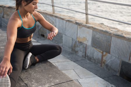 A woman in tights and top looking at her smartwatch while doing exercises on the beach promenadeの写真素材