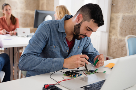 Man in a blue shirt with a beard is sitting at a desk and looking down at the pc board he is solderingの写真素材