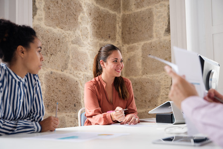 Two women and a man sitting at a desk smiling and talking in a room with a brick wall and two large windowsの写真素材